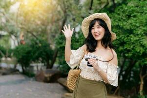 retrato de asiático joven mujer viajero con Costura sombrero y cesta y un cámara en verde público parque naturaleza antecedentes. viaje viaje estilo de vida, mundo viaje explorador o Asia verano turismo concepto. foto