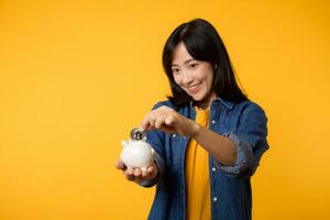 Happy young asian woman wearing yellow t-shirt denim shirt saving digital crypto currency to piggy bank isolated on yellow background. Digital money saving investment concept. photo
