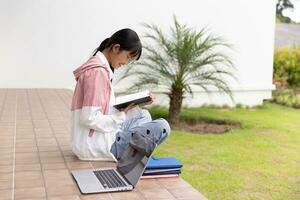 estudiante niña contento estudiando en línea, espalda a colegio concepto. joven mujer estudiante ocupado estudiar hacer notas en cuaderno y utilizando ordenador portátil concentrado en línea formación curso tecnología educación. foto