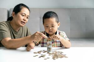 contento asiático niño y mamá ahorro dinero juntos, poniendo efectivo dentro cerdito banco. madre jugando con niño en calefacción piso a hogar, enseñando pequeño hijo a invertir dinero, planificación futuro. foto