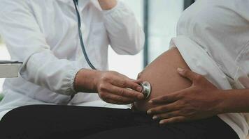 pré-natal saúde Cuidado. fechar acima do jovem grávida mulher visitando médico às pré-natal clínica. médico ginecologista examinar fetal batimento cardiaco taxa comovente estetoscópio de nu bebê colisão do expectante mãe video