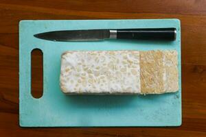 Fresh raw tempeh, on a cutting board and knife for chopping. Food preparation concept photo