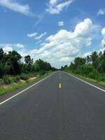 Image of new paved road in the daytime in the countryside painted yellow dividing line with trees, grass along the road, sky with white clouds photo