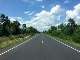 imagen de nuevo pavimentado la carretera en el tiempo de día en el campo pintado amarillo divisor línea con árboles, césped a lo largo el camino, cielo con blanco nubes foto