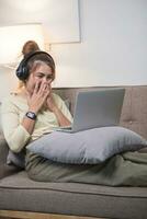 Young beautiful asian woman relaxing and listening to music using headphones, she is lying in sofa. Young woman enjoying at home using laptop. photo