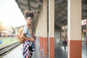 Young Asian African woman traveler with backpack in the railway train station, traveler girl walking stand sit waiting take a picture on railway platform train station. High quality photo