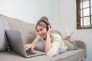 Young beautiful asian woman relaxing and listening to music using headphones, she is lying in sofa. Young woman enjoying at home using laptop. photo