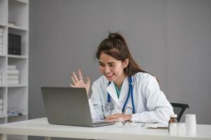 Young Asia lady doctor in white medical uniform with stethoscope using computer laptop talking video conference call with patient at desk in health clinic or hospital. Consulting and therapy concept. photo