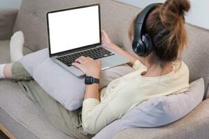 Young beautiful asian woman relaxing and listening to music using headphones, she is lying in sofa. Young woman enjoying at home using laptop. photo