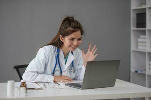 Young Asia lady doctor in white medical uniform with stethoscope using computer laptop talking video conference call with patient at desk in health clinic or hospital. Consulting and therapy concept. photo