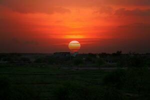 the sun sets over a field with a small village in the background photo