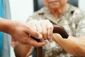 Doctor help Asian elderly disability woman patient holding walking stick in wrinkled hand at hospital. photo