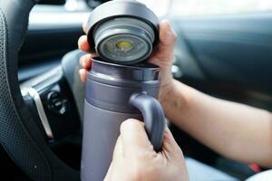 Asian woman driver hold thermo mug with hot coffee in car, dangerous and risk an accident. photo