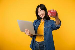 Happy young woman of Asian ethnicity wear yellow t-shirt denim shirt using laptop pc computer hold credit bank card shopping online order delivery isolated on plain yellow background. photo