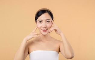 Beautiful girl asian model touching fresh glowing hydrated facial skin on beige background closeup. Beauty face young woman with natural makeup and healthy skin portrait. Skin care concept photo