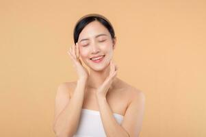 Beautiful girl asian model touching fresh glowing hydrated facial skin on beige background closeup. Beauty face young woman with natural makeup and healthy skin portrait. Skin care concept photo