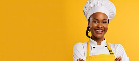 Joyful African American female cook in uniform and hat happily glancing at oven mitt on hand posing with delight in yellow studio backdrop photo