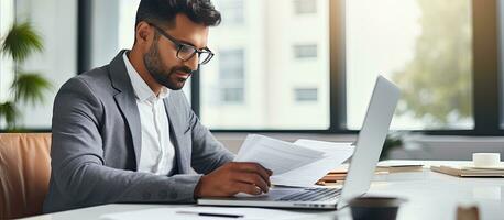 Concentrated businessman from Hispanic background working in office using laptop for online market research while holding documents photo