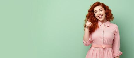 Cheerful young redhead woman in a pink dress pointing at empty space on green background photo