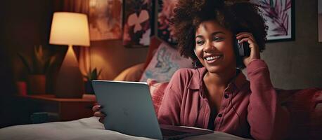 Happy African American woman multitasking on bed using laptop and cellphone for remote work or online shopping photo