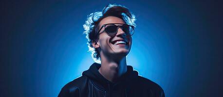 A fashionable teenage boy wearing a black longsleeve and glasses poses with a smile against a blue background illuminated by neon lights representing a hi photo