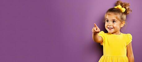 Cheerful child in yellow dress happily points at copyspace on purple background photo