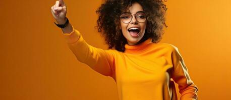 Enthusiastic young woman playfully imitates a rider dances energetically and happily points upwards dressed casually posing in a studio with an orange bac photo