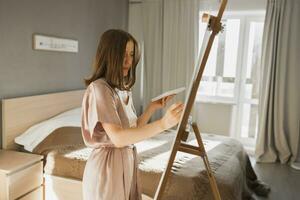 Young woman artist painting on canvas on the easel at home in bedroom - art and creativity concept photo