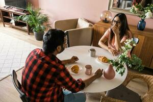 Happy couple eating breakfast and talking at dining table in morning. Indian girl and latino guy. Relationship and diversity concept photo