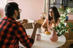 Happy couple eating breakfast and talking at dining table in morning. Indian girl and latino guy. Relationship and diversity concept photo