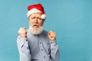 Happy confident cool old bearded Santa Claus winner raising fists celebrating triumph and success over blue background with copy space photo