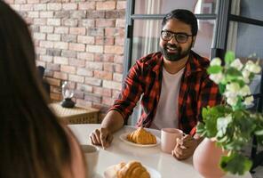 Happy couple eating breakfast and talking at dining table in morning. Indian girl and latino guy. Relationship and diversity concept photo