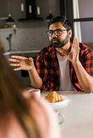 Happy indian couple having breakfast and small talk together in the kitchen - friendship, dating and family photo