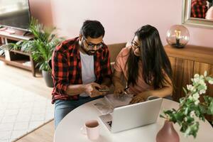 milenario indio marido y esposa haciendo Doméstico papeleo, contabilidad trabajo y revisando papel facturas, ingresos a ordenador portátil computadora, utilizando en línea calculadora - económico crisis concepto foto