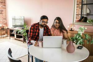 Indian family couple cuddle at desk make video call to friends using laptop webcam. Loving young spouses look at computer screen waving hands  communicating online via app photo