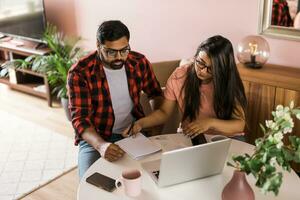 milenario indio marido y esposa haciendo Doméstico papeleo, contabilidad trabajo y revisando papel facturas, ingresos a ordenador portátil computadora, utilizando en línea calculadora - económico crisis concepto foto