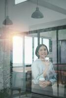 Happy young Asian saleswoman looking out the window. Smiling woman executive manager, secretary offering professional business services holding cup of coffee in office. Portrait photo