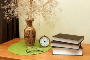 books with glasses and an alarm clock on the table next to a vase with a bouquet photo