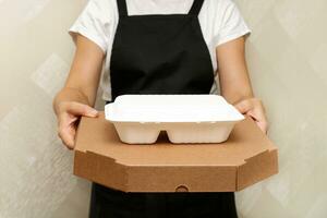 un mujer café trabajador sirve un terminado para llevar orden - Pizza en un caja y un envase con comida en desechable utensilios hecho desde reciclado crudo materiales foto