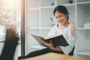 Side view of asian woman sitting armchair with open book and reading in living room. photo