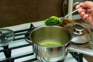 mano sostiene un cuchara con brócoli terminado un cacerola en el estufa foto