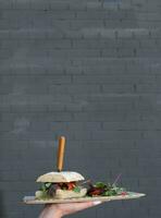 Waitress hand holding a burger with meat on brik wall background photo