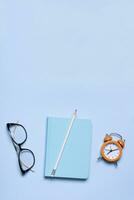 Notebook, glasses and alarm clock on blue background with copy space. Business minimalist concept photo
