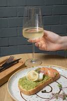 vaso de blanco vino en hembra mano y brindis con salmón, aguacate y escalfado huevo foto