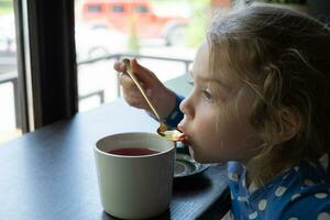 un pequeño niña bebidas té con un cucharilla en un café foto