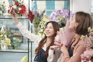 comercio electrónico negocio pareja, dos joven hembra florista trabajadores espectáculo floral acuerdo, en línea En Vivo transmisión, y selfies con teléfono inteligente solicitud en brillante flor comercio, hermosa flores almacenar. foto