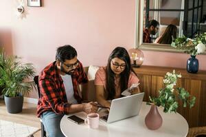 milenario indio marido y esposa haciendo Doméstico papeleo, contabilidad trabajo y revisando papel facturas, ingresos a ordenador portátil computadora, utilizando en línea calculadora - económico crisis concepto foto