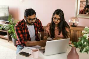 millennial indian husband and wife doing domestic paperwork, accounting job and reviewing paper bills, receipts at laptop computer, using online calculator - economic crisis concept photo