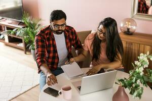 millennial indian husband and wife doing domestic paperwork, accounting job and reviewing paper bills, receipts at laptop computer, using online calculator - economic crisis concept photo