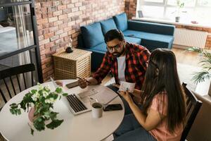 milenario indio marido y esposa haciendo Doméstico papeleo, contabilidad trabajo y revisando papel facturas, ingresos a ordenador portátil computadora, utilizando en línea calculadora - económico crisis concepto foto